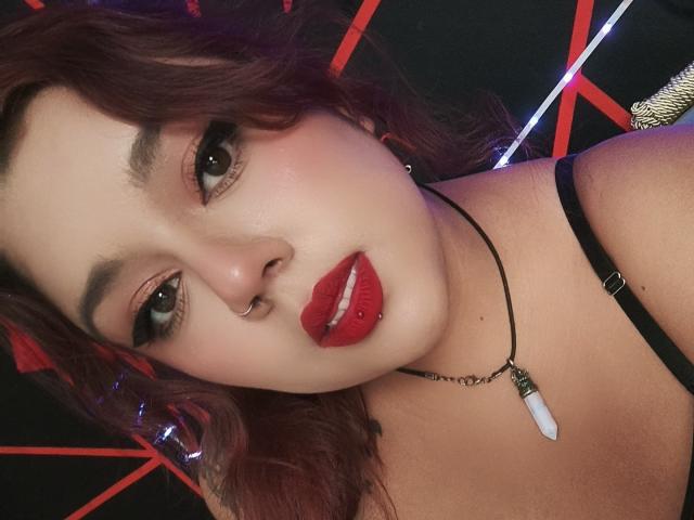 EmilyDom has red lipstick, a nose ring, and wavy brown hair. She's wearing a black strap and a necklace with a white stone. Her tits are partially visible.