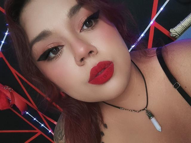 EmilyDom with red lipstick, black eyeliner, and nose ring, wearing a black strap, necklace with white stone, and red hair, looking at camera with slightly pouty lips.