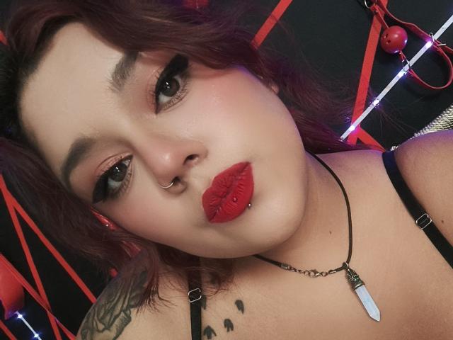 EmilyDom has red lipstick, a nose piercing, and a lip piercing, with dark makeup and red hair, wearing a black strap top and a necklace with a white stone.