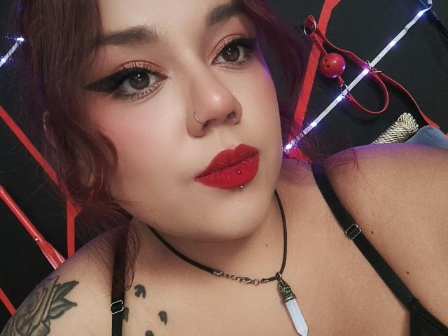 EmilyDom with red lipstick, eyeliner, and nose ring, has a lip piercing and tattoo on her shoulder, wearing black straps and a necklace, with a black background.