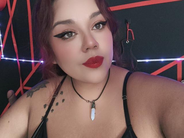 EmilyDom with red lipstick, black eyeliner, and a nose ring, wearing a black top and necklace with a white stone, has a tattoo on her left shoulder, looking at camera.