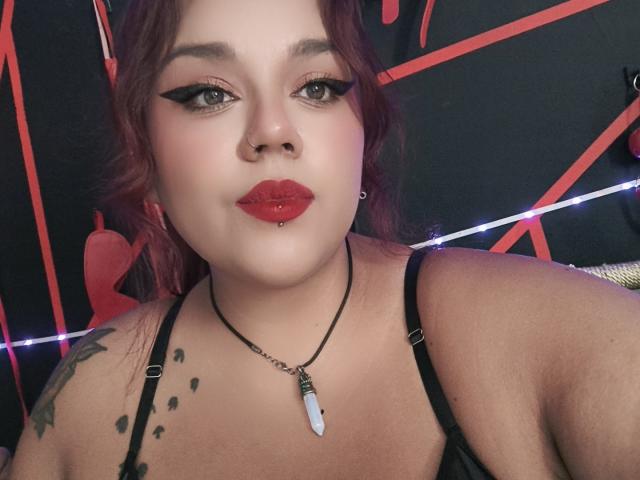 EmilyDom with red lipstick, black eyeliner, nose ring, and a black necklace with a white stone, showing tattoos on her right shoulder. She's wearing a black top.
