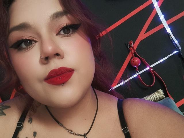 EmilyDom with red lipstick, pierced lip, nose ring, black eyeliner, and red eyeshadow, wearing a black strap top, showing tattoos, and a chain necklace.