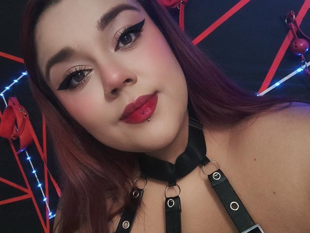 EmilyDom with red lipstick, black eyeliner, and a nose piercing, wearing a black leather harness, has a lip piercing, and red hair, smiling at the camera.