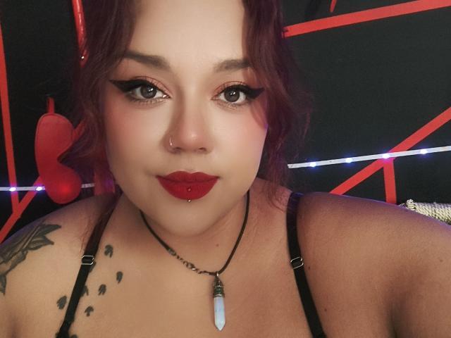 EmilyDom with red lipstick, black eyeliner, nose ring, and pierced lips, wearing a black strap top, necklace with a white stone, and tattoos on her shoulder.