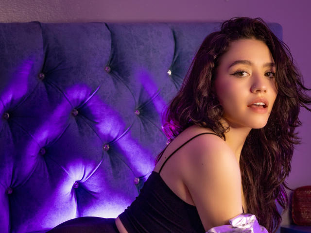 KateCrown with wavy dark brown hair, black crop top, and off-shoulder white top, looking at camera, purple-tufted headboard with lights in background. Soft purple light on headboard.