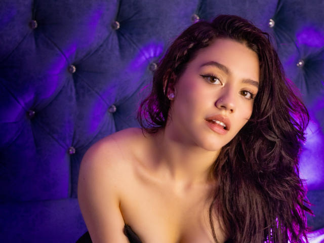 KateCrown has wavy dark brown hair, light brown skin, and is looking at the camera with pink lips, wearing earrings, and a black strapless top, against a purple tufted background.
