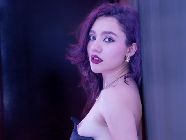 KateCrown with purple hair, dark lipstick, and gold earrings, leaning against a wall, showing her bare tits, looking over her shoulder, tattoo on her neck, purple-blue lighting.