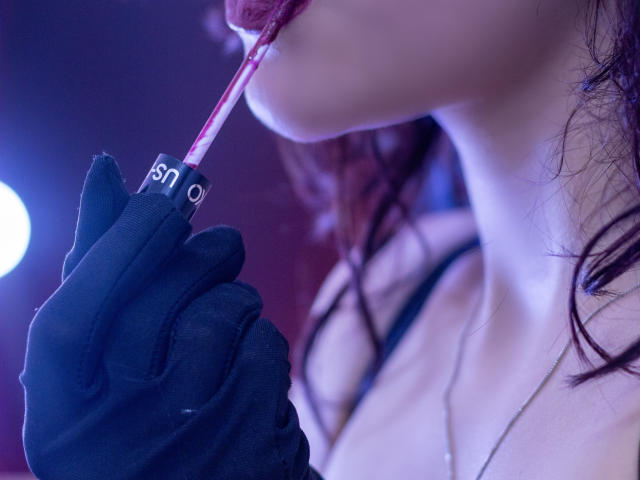 KateCrown's mouth is slightly open, wearing black gloves, holding a pink-and-white striped cigarette near her lips. Her red hair is visible, and she's wearing a black top.