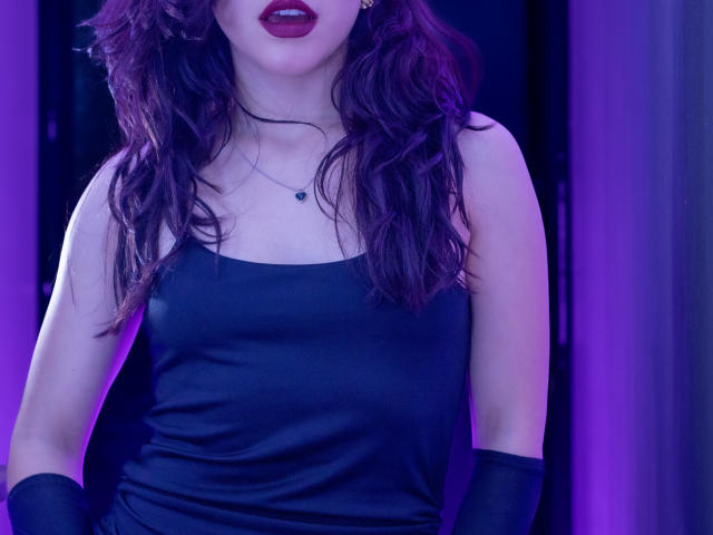 KateCrown with wavy purple hair, dark red lipstick, black sleeveless dress, and black gloves, showing her boobs and a small necklace. Purple and blue lighting.