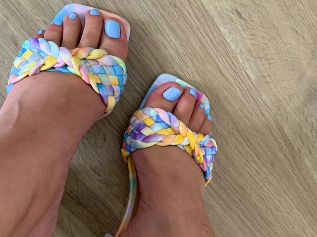 Les pieds de Babycamy-hot, ornés de sandales colorées tissées avec des lanières bleu pastel, jaune et violet, et des ongles des orteils peints en bleu assorti, se tiennent sur un plancher en bois..