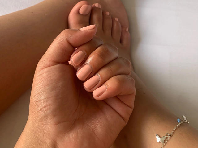 Babycamy-hot tient ses orteils avec sa main, ses ongles roses étant manucurés. Elle porte un bracelet en argent avec une petite pierre bleue. Sa peau est bronzée, et l'attention est portée sur sa main..