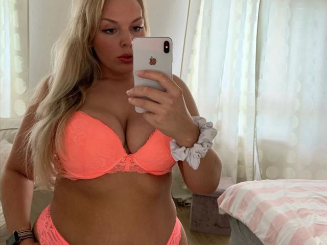 Babycamy – heiß, mit blonden Haaren, trägt einen neonrosa Spitzen-BH und passende Unterwäsche, hält ein iPhone, zeigt ihre Brüste und ein weißes Haargummi an ihrem Handgelenk..