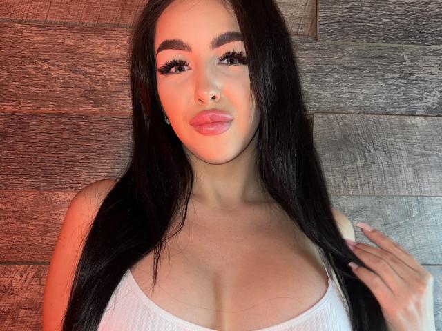 xJuliax has long black hair, full eyebrows, long lashes, pink lips, and big tits in a white top, holding her hair with a cute smile.