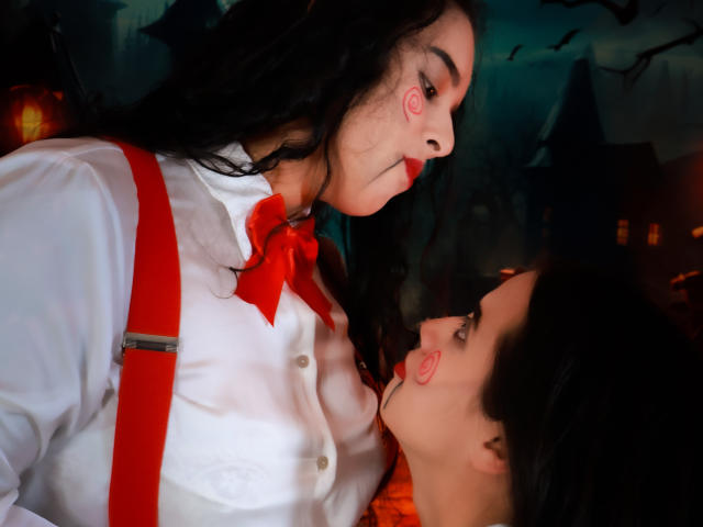 DilanAndMia, con cabello negro largo, camisa blanca, corbata roja y tirantes rojos, se inclina, con la nariz cerca de la de Mia, quien tiene cabello negro y lápiz labial rojo, y mira hacia arriba..