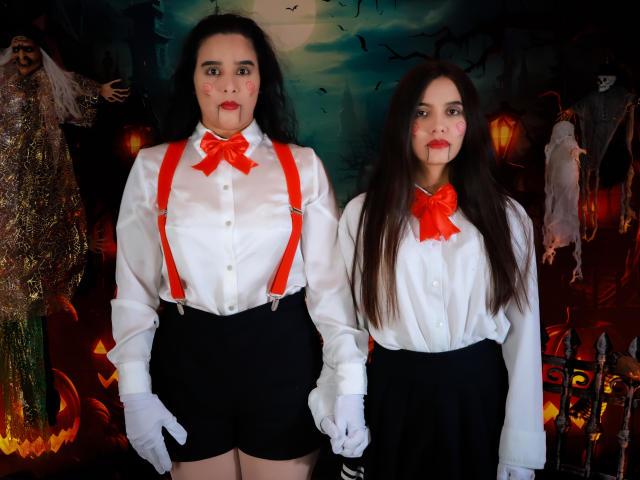 DilanAndMia con cabello oscuro, camisas blancas, lazos rojos, pantalones cortos negros y guantes blancos, de pie, tomadas de la mano, con tirantes rojos, sangre falsa en los rostros, guantes blancos, senos medianos..