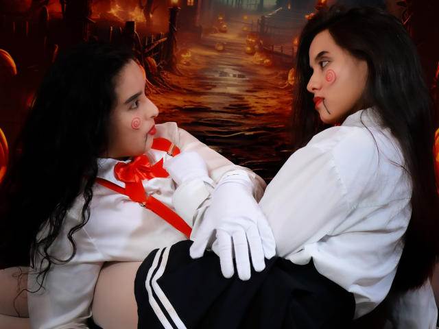 Dilan y Mia, con cabello negro largo, camisas blancas, lazos rojos y faldas negras, se enfrentan, tocándose las manos, usando guantes blancos, con pequeñas manchas rojas en las mejillas, sentadas en una luz tenue..