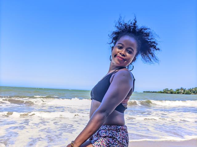Eme with dark skin, curly black hair, smiling, wearing a black sports bra and floral skirt, standing on a sandy beach with blue water and clear sky.
