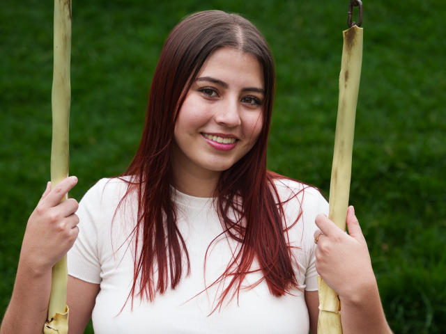 Georgia Gauthier houdt bamboestokjes vast, ze glimlacht met haar bruine haar met rode punten, een wit T-shirt en een achtergrond van groen gras. Ze heeft mooie borsten en een mooi gezicht..