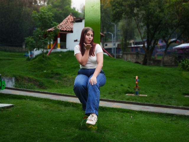 GeorgiaGauthier is sitting on a green pole, wearing a white shirt, blue jeans, and white sneakers, with her hand near her mouth, looking thoughtful. Her ass is slightly visible.