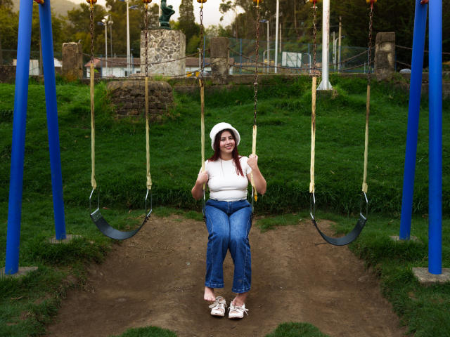 GeorgiaGauthier is sitting on a swing with blue poles, wearing a white hat, white shirt, blue jeans, and white sandals. She has tits and is smiling.