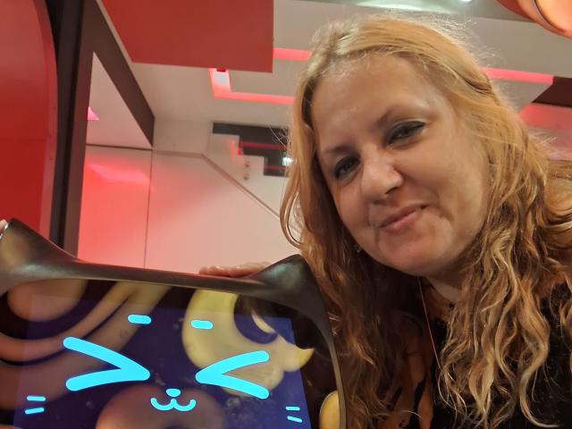 AmantsChauds ha i capelli biondi ondulati, la pelle chiara e un leggero sorriso, e tiene in mano una maschera di gatto luminosa e al neon con occhi blu. Indossa una maglietta nera..