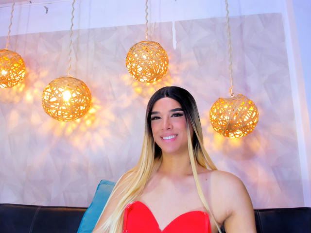 VictoriaAleia with long blonde hair, red strapless top, smiling, showing off her tits, hanging wicker lights, white wall, black couch, and blue pillow in the background.