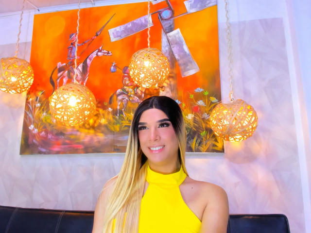 VictoriaAleia, con lunghi capelli biondi e neri, indossa un top giallo a halter, sorride ed è in piedi di fronte a un'opera d'arte arancione e gialla con tre lampade appese in vimini illuminate..