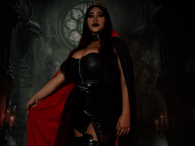 MelisaJackie with dark lipstick, black leather bustier, red cape, black skirt, and thigh-high boots, standing in a dark gothic-style room, showing her large, shiny, black-ass.