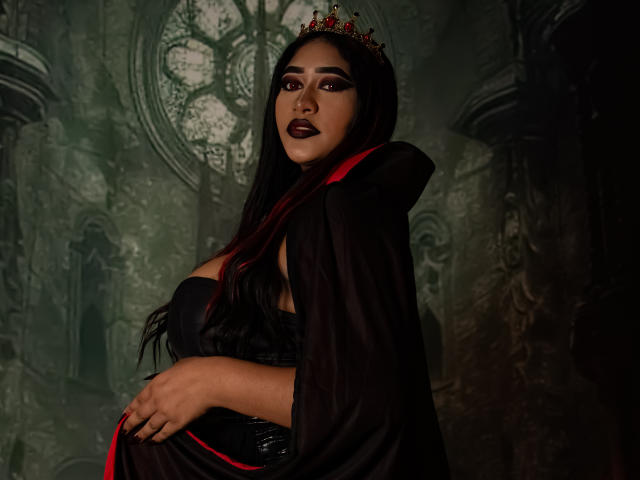 MelisaJackie with long black hair, dark lipstick, and a red crown, wearing a black cape over big boobs, standing with arms crossed, in a dark, Gothic-style setting.