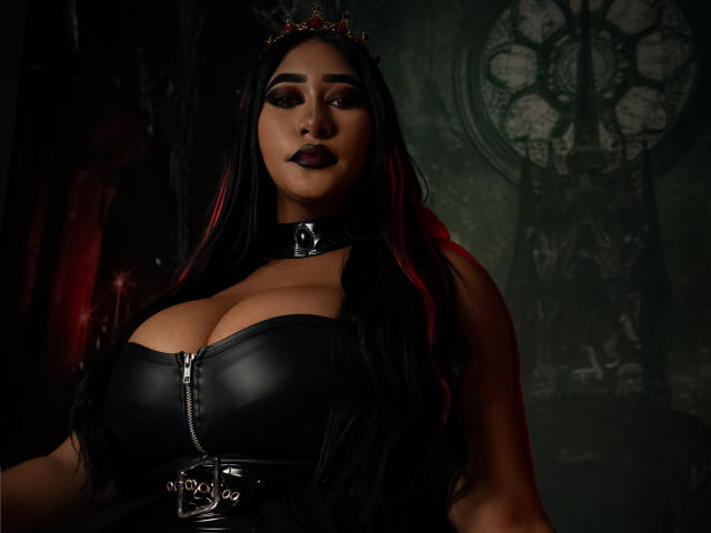 MelisaJackie with big boobs in a black leather bustier, zip down, wearing a shiny choker and crown, dark lipstick, long black hair, dark green background.
