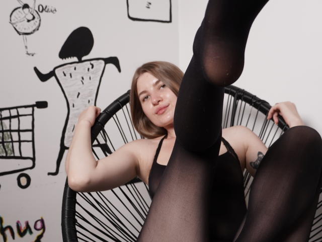 EllenMays with medium tits, black sheer pantyhose, and black top, legs up, in black wicker chair, smiling, light brown hair, tattoo on right arm, white wall background.