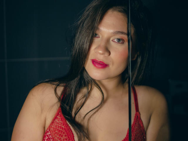 LindaJimenex, con cabello castaño largo, lápiz labial rojo y un sujetador de encaje rojo, está de pie frente a un panel de vidrio negro, mirando ligeramente hacia un lado con una sonrisa sutil..