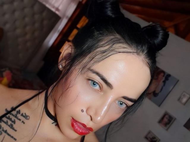 YeiSela with black double buns, blue eyes, red lipstick, and a small beauty mark on her left cheek, wearing a black choker, has a tattoo on her right shoulder.