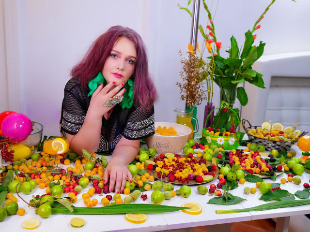 SinFulTitsDD with pink hair, green scarf, black mesh top, leans on table covered in colorful grapes, oranges, lemon slices, and flowers. SinFulTitsDD's hand touches mouth, looking cute.