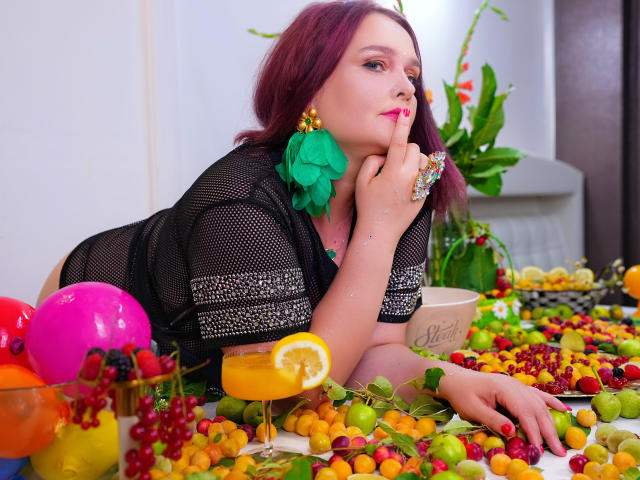 SinfulTitsDD with purple hair, black mesh top, green flower earrings, finger to lips, colorful fruit table, orange cocktail with lemon slice, pink balloon, white mug with "Cleed" in background.