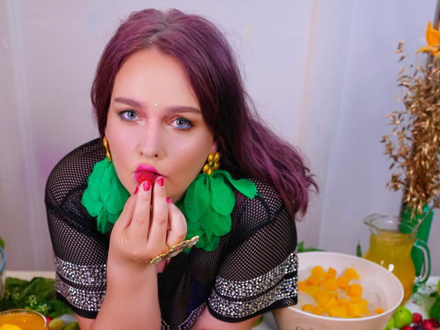 SinFulTitsDD with purple hair, wearing green leaf earrings, black mesh top, and pink lipstick, sucking on a red strawberry, hand with pink nail polish, yellow bracelet.