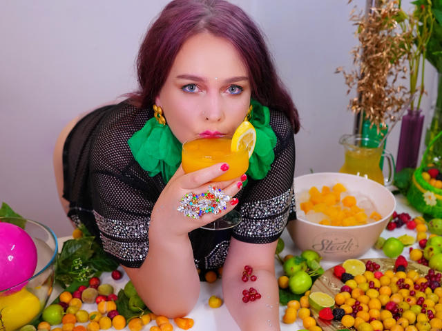 SinFulTitsDD with purple hair, wearing black mesh top and green collar, sucks orange drink through lemon slice, colorful nails, candy and fruit scattered, looking at camera.
