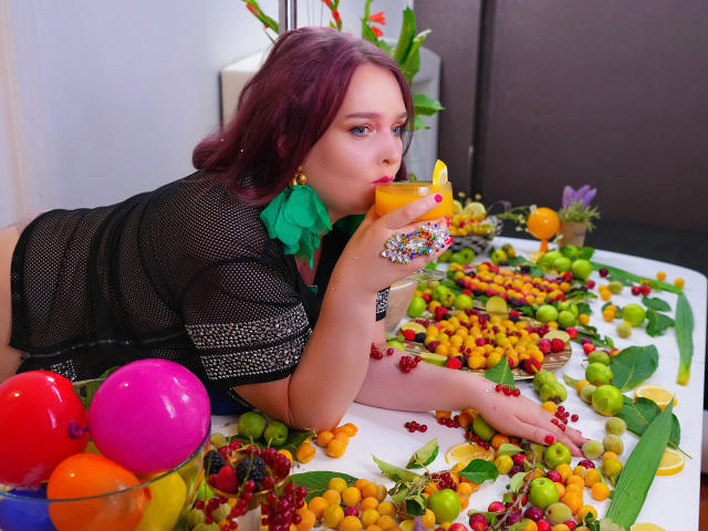 SinFulTitsDD with red hair, black mesh top, green bow, drinking orange cocktail, surrounded by colorful fruit, grapes, and balloons, lying on white table, wearing flower ring.