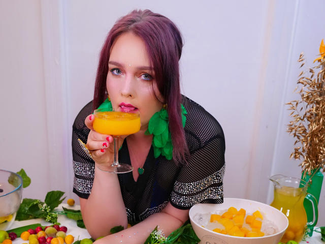 SinFulTitsDD with purple hair, green earrings, and black mesh top, sipping orange cocktail, holding glass to lips, colorful candies and orange slices on table, green bottle.