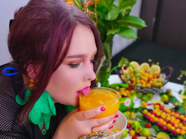 SinFulTitsDD with purple hair, green earrings, and pink lipstick, sips orange juice with tongue out, holding glass, surrounded by colorful fruit and green leaves.