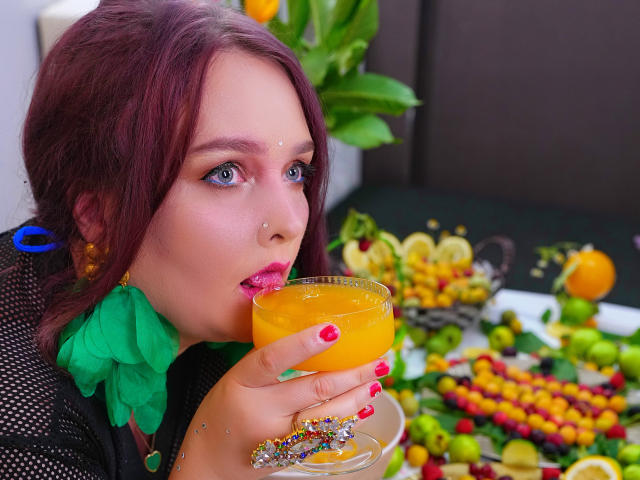 SinFulTitsDD with red hair, green ruffle collar, and blue earrings, sips orange cocktail, tongue out, pink lipstick, red nail polish, glitter on glass, colorful snack tray in background.