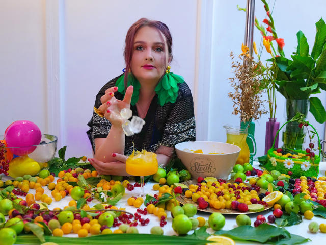 SinFulTitsDD with red hair, green earrings, and black mesh top, holding a white ruffle, snacking on grapes, surrounded by colorful fruits and a yellow drink, in a festive setup.