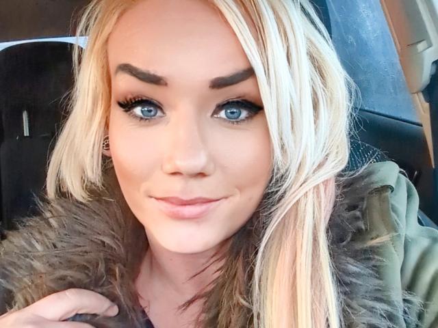 BustinBlonder with blonde hair, blue eyes, thick eyebrows, and a subtle smile, wearing a furry green jacket, sitting in a car. Her tits are not visible.
