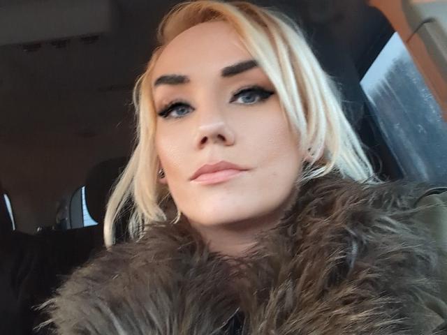 BustinBlonder has blonde hair, dark eyebrows, blue eyes, and pink lips, wearing a furry green coat. She's looking to the side with a slight pout.