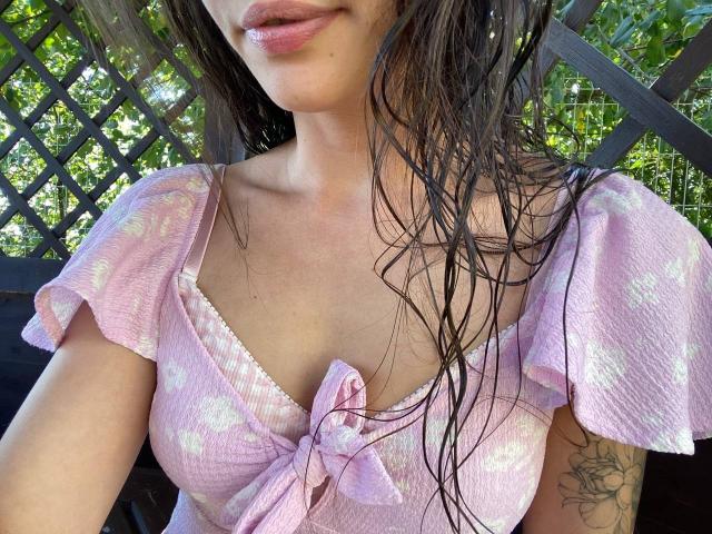 MoniFoxy with wet brown hair, pink ribbed top, tied below bust, small puffy sleeves, visible tattoo on right arm, slight smile, cute pink lips, soft natural light.