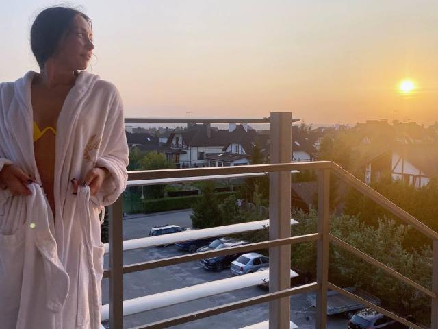 MoniFoxy in a white bathrobe, showing her tits in a yellow bra, stands on a balcony at sunset, holding the robe, looking right, with cars below.
