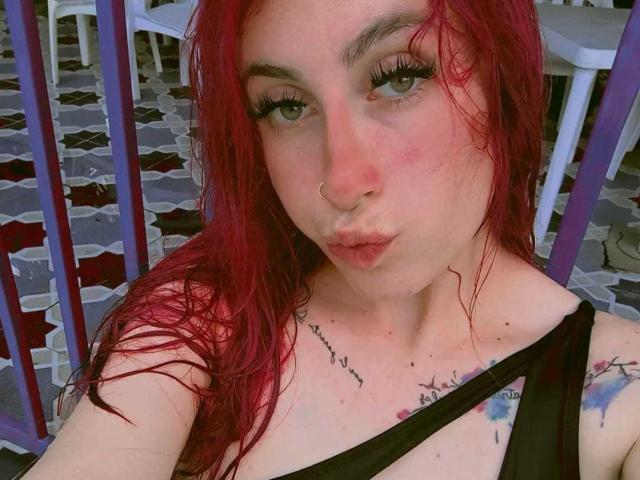 SammyViolet with red hair, nose ring, and tattoos, pouting lips, green eyes, black strap, and subtle makeup, showing her tits and tattoos, sitting outside.