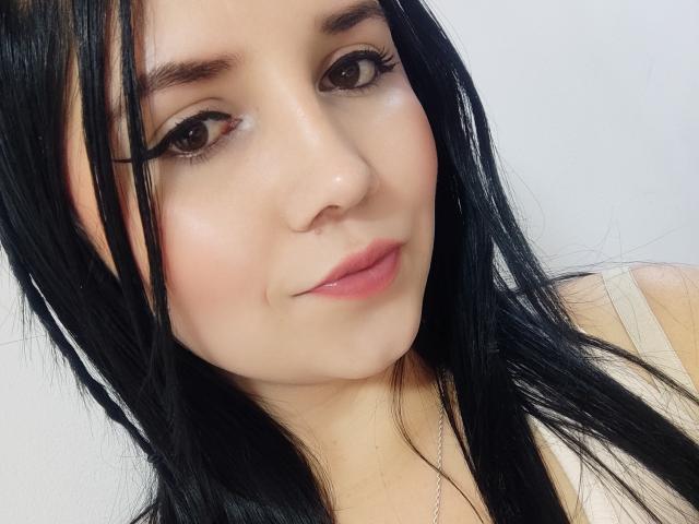 Kkatalina has long black hair, brown eyes, and pink lips with a slight smile. Her makeup includes eyeliner and she's wearing a white top. She's looking at the camera.