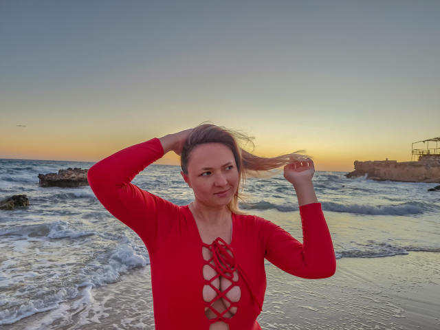 SallyVerda, con i capelli biondi, indossa una maglietta rossa con lacci, si tiene i capelli, sorride, è in piedi sulla spiaggia, ci sono onde, il tramonto, il cielo è sereno. Il seno è visibile attraverso la maglietta. Ha le mani tra i capelli. Aspetto casual, sicura di sé..